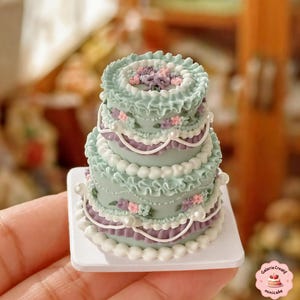 May include: A miniature three-tiered cake with a light teal frosting, decorated with white, purple, and pink details. The cake is presented on a small white square base, showcasing intricate details.