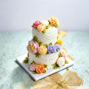 Puede incluir: Un pastel blanco de dos niveles decorado con flores de crema de mantequilla en tonos rosa, naranja, amarillo y morado, acentuado con hojas verdes. El pastel presenta un patrón de puntos texturizados en los lados y se asienta sobre una base cuadrada blanca.