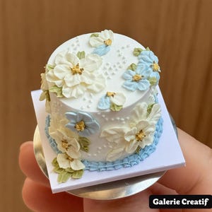 Puede incluir: Un pastel blanco en miniatura decorado con flores blancas y celeste, hojas verdes y detalles dorados. El pastel se asienta sobre una base cuadrada blanca con un borde rizado celeste y una base plateada. Es una versión en miniatura.