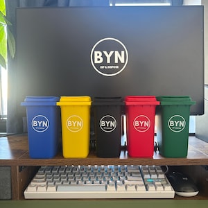 May include: A collection of five miniature trash cans in blue, yellow, black, red, and green. Each can has a white circular logo with the letters "BYN" and the words "DIP & DISPOSE". The cans are arranged in a row on a wooden surface.