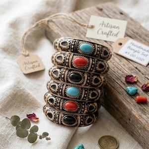 Op de afbeelding: Een stapel van zes donker metalen armbanden met ingewikkelde reliëfpatronen en centrale ovale cabochons in turkoois, rood en zwart. De armbanden gaan vergezeld van kleine papieren labels met de tekst Artisan Crafted en Handmade Bangle Set.