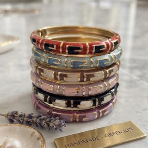 Op de afbeelding: Een stapel van zes scharnierende metalen armbanden met een herhalend Grieks sleutelpatroon in rood, lichtblauw, crème, roze, zwart en paars. Een rechthoekig goudkleurig metalen plaatje is gegraveerd met de tekst HANDMADE - GREEK KEY.