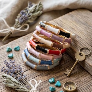 Op de afbeelding: Een stapel van zeven armbanden met een op bamboe geïnspireerd ontwerp en goudkleurige metalen accenten. Elke armband heeft een andere effen kleur, waaronder wit, lavendel, bruin, rood, zwart, roze, lichtblauw en grijs.