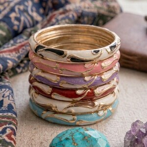 Puede incluir: Una pila de seis brazaletes circulares con una base de metal dorado e incrustaciones de esmalte de colores. Los brazaletes muestran varios colores, incluyendo blanco, rosa, morado, rojo y azul claro, con patrones de metal dorado.