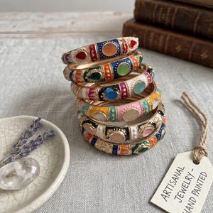 May include: A stack of six metal bangles featuring colourful enamel patterns and central oval stones. The bracelets display various hues including blue, orange, pink, and white. A rectangular tag reads ARTISANAL JEWELRY - HAND PAINTED.