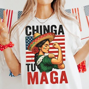 May include: White t-shirt with a graphic of a woman wearing a sombrero, flexing her arm in front of an American flag design. The text "CHINGA TU MAGA" is printed above and below the image.