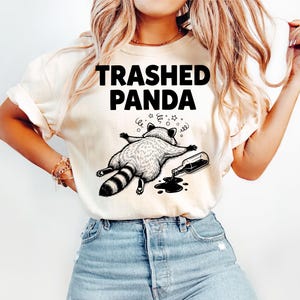 May include: A cream-coloured t-shirt featuring the text "Trashed Panda" and a black and white illustration of a raccoon lying down next to a spilled bottle. The raccoon has a striped tail and is surrounded by stars.