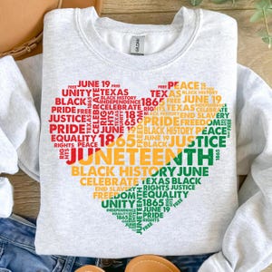 May include: A pale grey sweatshirt with a heart-shaped design made up of words in red, yellow, and green. The words include "Juneteenth," "Justice," "Freedom," and "Equality." The sweatshirt has a crew neck and long sleeves.