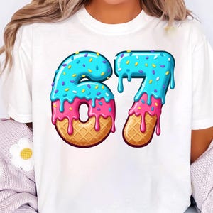 Pode incluir: Camiseta branca com o número 67 em um design de donut. Os números são azuis com granulado e cobertura rosa, sobre uma base de waffle. Uma camiseta gráfica colorida.