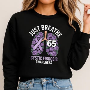 Puede incluir: Sudadera negra con un gráfico que promueve la concienciación sobre la fibrosis quística. El diseño incluye las palabras "Just Breathe" sobre una cinta morada y un diseño floral de pulmón con el número "65". Las palabras "Cystic Fibrosis Awareness" están debajo.