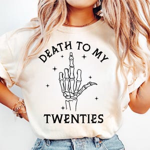 May include: Cream-colored t-shirt with the text "DEATH TO MY TWENTIES" above a black skeleton hand giving the middle finger. The design includes small star accents.