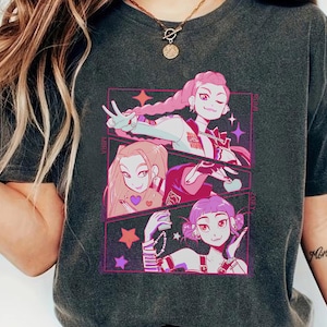 May include: Dark gray t-shirt featuring a colorful anime-style graphic of three female characters. The graphic is framed in a pink rectangle with stars. The characters have pink, brown, and purple hair.