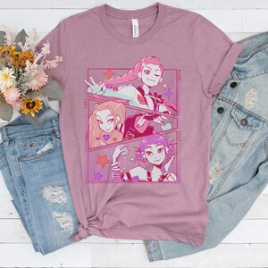 May include: Mauve-colored t-shirt featuring a manga-style illustration of three female characters. The artwork is framed in a pink border with star accents. The shirt is paired with light blue jeans and a denim jacket.
