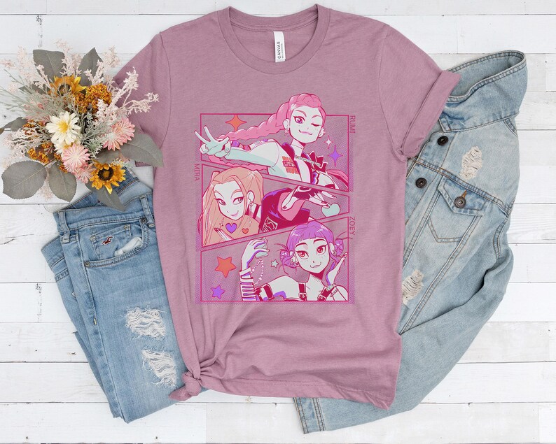 May include: Mauve-colored t-shirt featuring a manga-style illustration of three female characters. The artwork is framed in a pink border with star accents. The shirt is paired with light blue jeans and a denim jacket.
