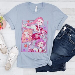 May include: Light blue t-shirt featuring a manga-style graphic with three female characters. The graphic includes the names Mira, Rumi, and Zoey. The shirt is paired with blue jeans and white sneakers.