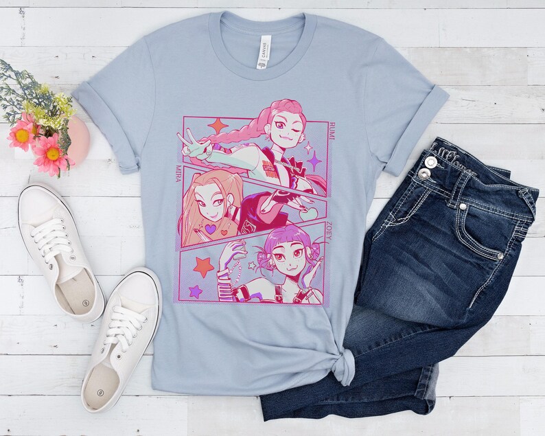 May include: Light blue t-shirt featuring a manga-style graphic with three female characters. The graphic includes the names Mira, Rumi, and Zoey. The shirt is paired with blue jeans and white sneakers.