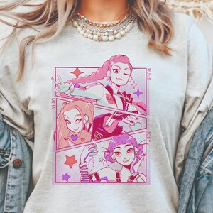 May include: A light gray t-shirt featuring a manga-style graphic with three female characters in a comic strip layout. The characters have pink, light brown, and purple hair. The names "MIRA", "RUMI", and "ZOEY" are printed on the side.