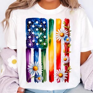 May include: White t-shirt featuring a watercolor design of a flag with rainbow stripes and white stars. The flag design is adorned with daisies and green leaves. The shirt is a casual, everyday wear item.