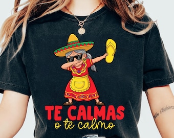 May include: Black t-shirt featuring a cartoon of a woman dabbing while wearing a sombrero, sunglasses, and a red dress with a yellow apron. The text "TE CALMAS o te calmo" is printed in red and yellow.
