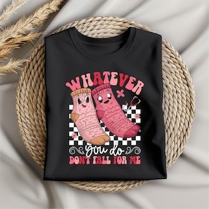 Puede incluir: Camiseta negra con un gráfico de dos calcetines rosas con caras, una venda y un estetoscopio. El texto "Whatever you do don't fall for me" está escrito debajo de los calcetines.