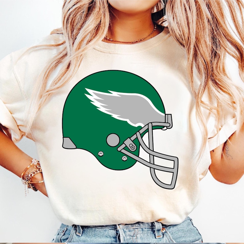 May include: White t-shirt with a green and gray football helmet graphic. The helmet has a wing design on the side.