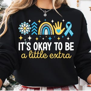 May include: Black sweatshirt with the text "IT'S OKAY TO BE a little extra" in white and yellow. The design includes a flower, trees, a rainbow, a hand with a heart, and a blue ribbon. The design is in blue, yellow, and white.