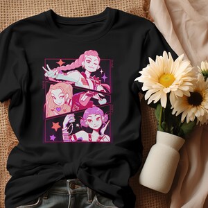 May include: Black t-shirt featuring a colorful anime-style graphic of three female characters. Each character is framed in a pink and purple rectangle with stars. The shirt is made of a soft material and is suitable for casual wear.