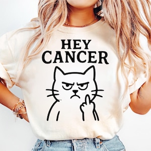 Może przedstawiać: Kremowa koszulka z czarnym napisem "HEY CANCER" nad czarną ilustracją kota z kreskówki. Kot pokazuje środkowy palec.