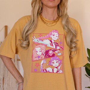 May include: Mustard yellow t-shirt featuring a colorful anime-style graphic. The graphic includes three panels with illustrations of anime characters. The text "MIRA" and "ZOEY" are visible. The shirt is a comfortable, casual style.