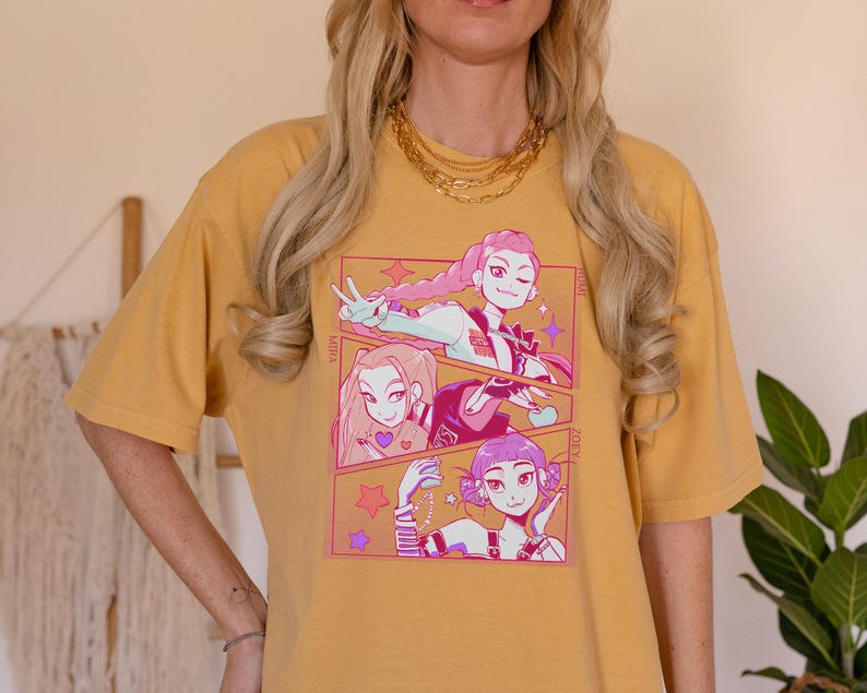 May include: Mustard yellow t-shirt featuring a colorful anime-style graphic. The graphic includes three panels with illustrations of anime characters. The text "MIRA" and "ZOEY" are visible. The shirt is a comfortable, casual style.