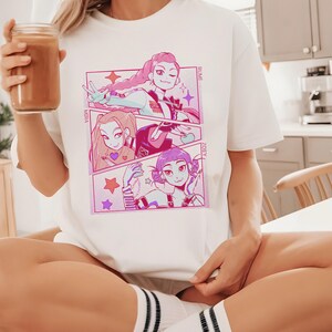 May include: White t-shirt featuring a colorful anime-style graphic. The graphic is divided into three panels, each showcasing a different character with pink and purple accents. The names "MIHA", "RUMI", and "ZOEY" are printed on the shirt.