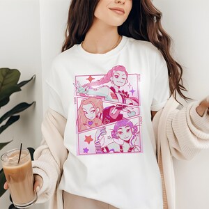 May include: White t-shirt featuring a manga-style graphic in pink and purple. The design is divided into three panels, each showcasing a different character with text labels. The shirt is a casual, everyday wear item.