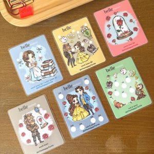 Può includere: Una collezione di carte Belle Minis con illustrazioni di personaggi e icone de La Bella e la Bestia. Ogni carta ha un design unico con la scritta "belle minis". Le carte sono su una superficie di legno.