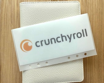 May include: A white textured wallet with a clear plastic sleeve featuring the "Crunchyroll" logo in gray and orange. The wallet has a gold ring and pearl charm attached to the zipper.