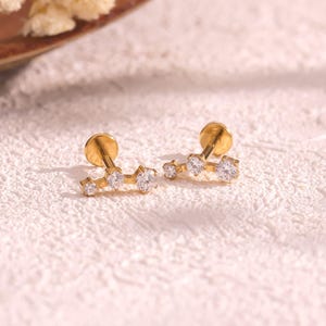 May include: A pair of gold stud earrings with a constellation design. Each earring has a small, round post and a curved bar with three clear, sparkling gemstones. The earrings are set against a textured white surface.