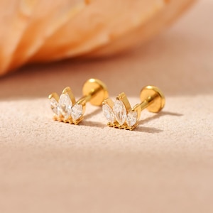 May include: Two gold-colored stud earrings with a crown design. Each earring features three clear, marquise-cut gemstones. The earrings are displayed on a light-colored surface, with a blurred orange background.