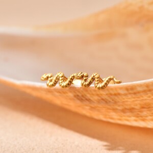 K&ouml;nnte beinhalten: Ein goldfarbener, schlangenf&ouml;rmiger Ohrring. Der Ohrring ist ein kleines, filigranes Schmuckst&uuml;ck mit strukturierter Oberfl&auml;che. Das Schlangenmuster ist gewunden und detailliert, auf einer hellbraunen Oberfl&auml;che liegend.