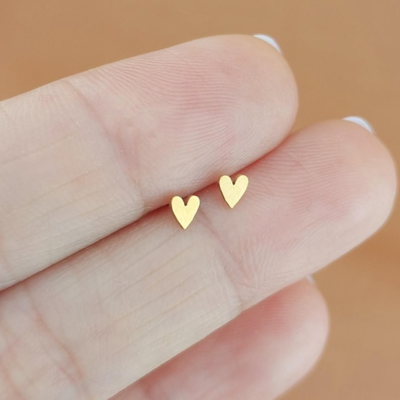 May include: A pair of small, gold-colored heart-shaped stud earrings. The earrings are simple and minimalist in design, with a smooth, polished finish. The earrings are shown on a person's finger.