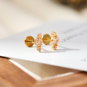 May include: A pair of gold-tone stud earrings featuring a central round peach-colored stone flanked by two smaller matching stones on each side. The earrings have a flat circular backing and a post mechanism for secure attachment to the earlobe.