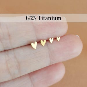 May include: Four gold-colored heart-shaped stud earrings in graduated sizes. The earrings are displayed on a finger, with the text "G23 Titanium" above them. The background is a warm, neutral tone.
