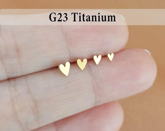 May include: Four gold-colored heart-shaped stud earrings in graduated sizes. The earrings are displayed on a finger, with the text "G23 Titanium" above them. The background is a warm, neutral tone.