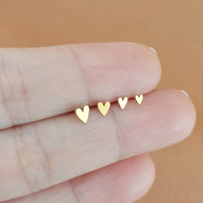 May include: Four small, gold-colored heart-shaped stud earrings. The earrings are arranged in a row, decreasing in size from left to right. The earrings are displayed on a person's finger.