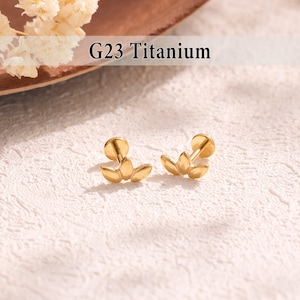 May include: Two gold-colored G23 Titanium earrings. Each earring features a three-petal design. The earrings are displayed on a textured, off-white surface. The text "G23 Titanium" is visible above the earrings.