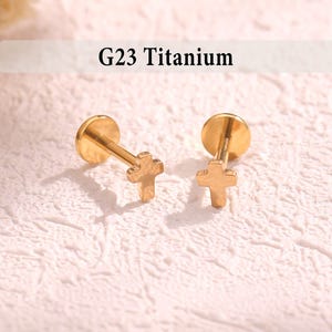 May include: Two gold-colored G23 Titanium earrings. Each earring features a cross-shaped charm attached to a post with a flat disc backing. The earrings are displayed on a textured, off-white surface.
