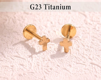 May include: Two gold-colored G23 Titanium earrings. Each earring features a cross-shaped charm attached to a post with a flat disc backing. The earrings are displayed on a textured, off-white surface.