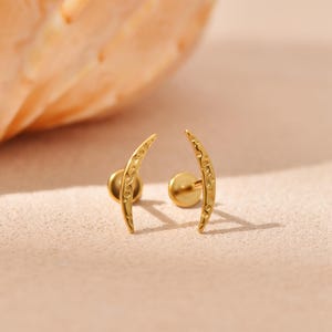 May include: A pair of gold-toned stud earrings featuring a curved, crescent moon shape. The surface of each earring has a textured, hammered finish with small indentations. Each earring includes a flat circular backing post for secure attachment.