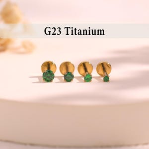 May include: Four gold-colored titanium earrings with emerald green gemstones. The earrings are arranged in a row, with the text "G23 Titanium" above them.