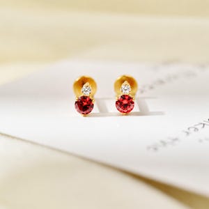 May include: A pair of gold stud earrings, each featuring a round, faceted red gemstone set below a smaller, clear, round, faceted gemstone. The earrings are arranged side-by-side, showcasing a bright, polished gold metal setting for each stone.