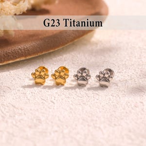 May include: Four small paw-shaped earrings. Two are gold-colored, and two are silver-colored. The earrings are made of G23 Titanium. The text "G23 Titanium" is displayed in the image.