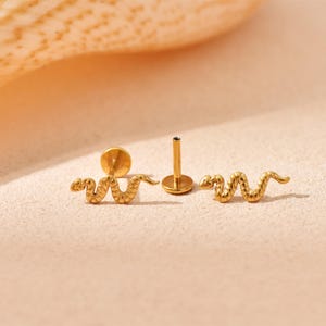 K&ouml;nnte beinhalten: Goldfarbene Ohrringe in Schlangenform. Die Ohrringe haben eine strukturierte Oberfl&auml;che und ein Stecker-Design. Das Bild zeigt zwei Schlangenohrringe und einen einzelnen Ohrringpfosten. Die Ohrringe werden auf einer hellrosa Oberfl&auml;che pr&auml;sentiert.